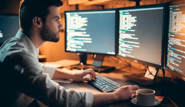 Focused young developer coding late in office writing script shown on computer monitors, serious handsome coder programmer hacker programming developing software applications working alone at night