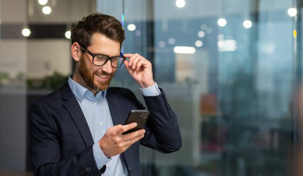 Successful financier investor works inside office at work, businessman in business suit uses telephone near window, man smiles and reads good news online from smartphone.