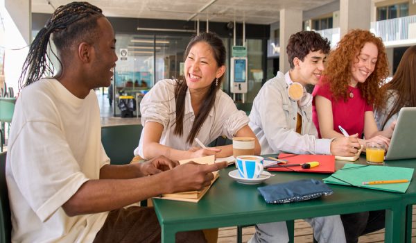 Group of cheerful multicultural vocational students gathered in high school cafeteria to homework and discuss exam topic. Young multiracial classmates using laptop and taking notes in notebooks.