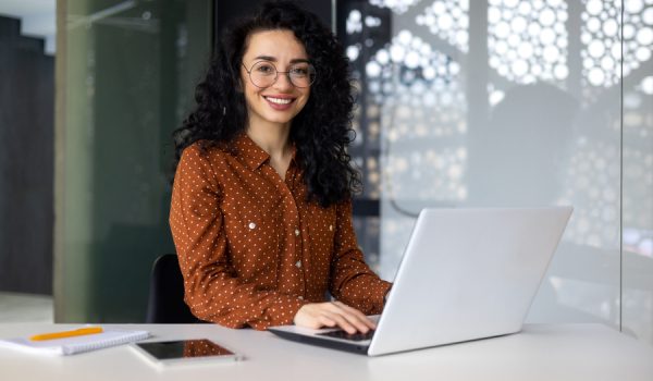 Happy and smiling hispanic businesswoman typing on laptop, office worker with curly hair and glasses happy with achievement results, at work in office building, portrait of female boss looking at camera.